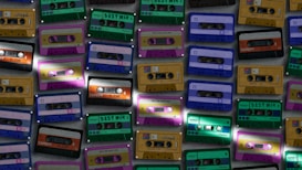 Various cassette tapes in different colors are scattered, showcasing vibrant designs and labels. The tapes are arranged in an overlapping pattern, creating a retro and nostalgic feel.