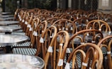 Numerous outdoor bistro chairs and tables are stacked closely together, suggesting an arrangement typical of a cafe or restaurant setting. The chairs have curved wooden frames and woven seats, while the round tables have smooth tops.