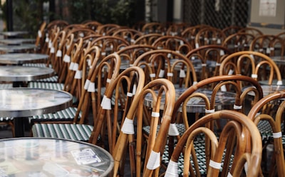 Numerous outdoor bistro chairs and tables are stacked closely together, suggesting an arrangement typical of a cafe or restaurant setting. The chairs have curved wooden frames and woven seats, while the round tables have smooth tops.