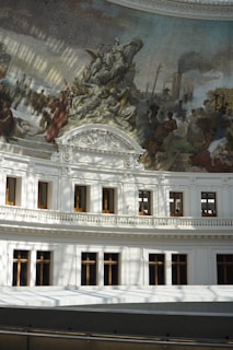 A grandiose interior featuring a large mural painted on a domed ceiling. The mural showcases various historical and allegorical figures engaged in different activities. Below the mural, there are intricately detailed white architectural elements including arched windows and elaborate cornices. The light coming through casts shadows across the scene.