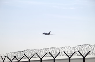 A distant airplane is flying in a clear sky above a barbed wire fence. The fence is topped with spiral coils of barbed wire, suggesting a secure or restricted area.