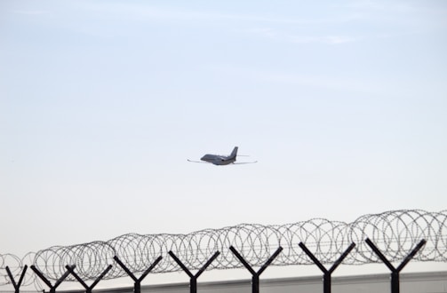 A distant airplane is flying in a clear sky above a barbed wire fence. The fence is topped with spiral coils of barbed wire, suggesting a secure or restricted area.