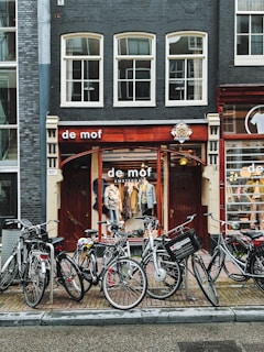A charming street scene in Amsterdam with a vintage storefront named 'de mof'. Multiple bicycles are parked in front of the shop, which has a display of clothing inside visible through large windows with a wooden frame. The building facade is characterized by dark bricks and large, rectangular windows.