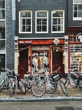 A charming street scene in Amsterdam with a vintage storefront named 'de mof'. Multiple bicycles are parked in front of the shop, which has a display of clothing inside visible through large windows with a wooden frame. The building facade is characterized by dark bricks and large, rectangular windows.