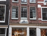 A storefront with a red brick facade and large black-framed windows. The shop is named 'Fleurmonde' as indicated by the illuminated sign above the entrance. The exterior has white decorative elements and there is an architectural relief above the windows depicting a fish or sea creature. Inside, stylish light fixtures are visible.