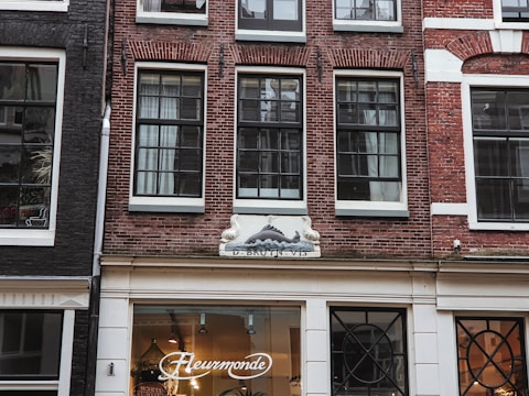 A storefront with a red brick facade and large black-framed windows. The shop is named 'Fleurmonde' as indicated by the illuminated sign above the entrance. The exterior has white decorative elements and there is an architectural relief above the windows depicting a fish or sea creature. Inside, stylish light fixtures are visible.