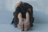 A person with long, dark curly hair is sitting on a textured surface, stretching forward with their hands reaching for their bare feet. The setting is softly lit, giving a sense of calm and focus.