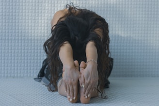 A person with long, dark curly hair is sitting on a textured surface, stretching forward with their hands reaching for their bare feet. The setting is softly lit, giving a sense of calm and focus.