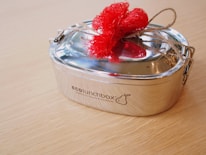 A stainless steel lunchbox with a polished finish is tied with twine and adorned with a bright red fabric bow, placed on a light wooden surface.