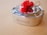 A stainless steel lunchbox with a polished finish is tied with twine and adorned with a bright red fabric bow, placed on a light wooden surface.
