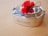 A stainless steel lunchbox with a polished finish is tied with twine and adorned with a bright red fabric bow, placed on a light wooden surface.