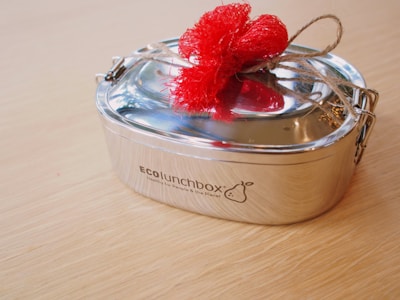 A stainless steel lunchbox with a polished finish is tied with twine and adorned with a bright red fabric bow, placed on a light wooden surface.