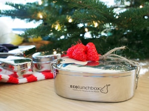 A shiny metal lunchbox with a red decorative item on top is placed on a wooden surface. In the background, a lush green Christmas tree with lights is visible, creating a festive atmosphere. Additional metal containers and a red and white striped cloth are placed nearby.