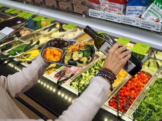 A person is holding a metal plate with a variety of fresh salad ingredients, including roasted vegetables and greens. They are about to pour olive oil from a bottle onto the salad. In the background, there is a salad bar with various fresh ingredients such as cherry tomatoes, chopped broccoli, and a variety of pickles.