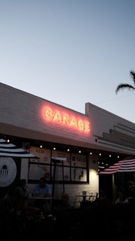 A pastel sky provides the backdrop for a brick building adorned with a vibrant red neon sign reading 'GARAGE'. Below, a cozy outdoor seating area features striped umbrellas and dim lighting, hinting at a casual dining atmosphere. The area is populated with people enjoying their meals at small tables. A palm tree is visible in the background, adding to the laid-back vibe.