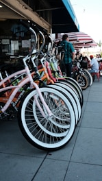 A row of colorful bicycles is neatly parked on a sidewalk in front of a shop. The bicycles have curved handlebars and bright frames, including pink, orange, and green. A person is standing nearby, and behind them, there are tables with people sitting under red and white striped umbrellas.
