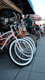 A row of colorful bicycles is neatly parked on a sidewalk in front of a shop. The bicycles have curved handlebars and bright frames, including pink, orange, and green. A person is standing nearby, and behind them, there are tables with people sitting under red and white striped umbrellas.