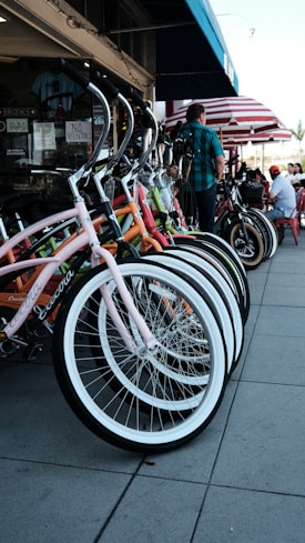 A row of colorful bicycles is neatly parked on a sidewalk in front of a shop. The bicycles have curved handlebars and bright frames, including pink, orange, and green. A person is standing nearby, and behind them, there are tables with people sitting under red and white striped umbrellas.