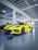 A bright yellow sports car with black racing stripes is parked inside a modern garage. The car has a sleek, aerodynamic design with visible black alloy wheels. In front of the car, a small black device with various components is placed on the garage floor. The garage itself is spacious, with a high ceiling, white walls, and exposed piping, creating an industrial look. Signs and office spaces can be seen in the background.