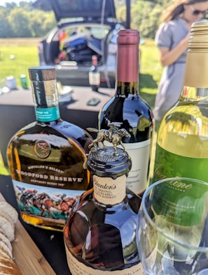 Several bottles of wine and whiskey are displayed on a table outdoors. In the background, a car with an open trunk is visible, along with a few items inside. A person stands on the right side, slightly out of focus.