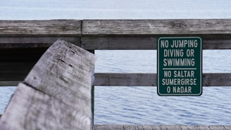 A wooden railing beside a body of water with a green warning sign attached. The sign contains white text in both English and Spanish, stating 'No Jumping, Diving or Swimming'. The water appears calm, and the scene suggests a peaceful, quiet environment.