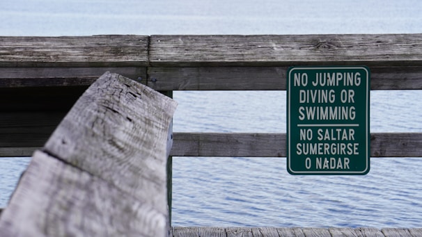 A wooden railing beside a body of water with a green warning sign attached. The sign contains white text in both English and Spanish, stating 'No Jumping, Diving or Swimming'. The water appears calm, and the scene suggests a peaceful, quiet environment.
