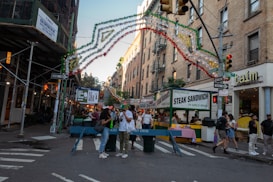 A lively street scene with festive decorations hanging over a busy intersection. Stalls selling food, like mozzarella steak sandwiches, are set up alongside the street. People are gathered around, some holding drinks, giving a vibrant and social atmosphere. The setting suggests a festival or street fair, with colorful garlands and food signs enhancing the environment.