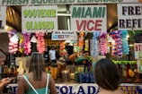 A busy stall at an outdoor event is decorated with colorful flower garlands and signs advertising fresh squeezed lemonade, souvenir glasses, and Miami Vice. Several people are standing in front of the stall, with one browsing their phone and another with their back to the camera. In the background, there are containers, cups, and a person preparing drinks.