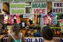 A busy stall at an outdoor event is decorated with colorful flower garlands and signs advertising fresh squeezed lemonade, souvenir glasses, and Miami Vice. Several people are standing in front of the stall, with one browsing their phone and another with their back to the camera. In the background, there are containers, cups, and a person preparing drinks.