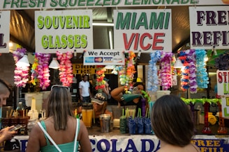 A busy stall at an outdoor event is decorated with colorful flower garlands and signs advertising fresh squeezed lemonade, souvenir glasses, and Miami Vice. Several people are standing in front of the stall, with one browsing their phone and another with their back to the camera. In the background, there are containers, cups, and a person preparing drinks.