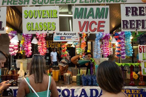 A busy stall at an outdoor event is decorated with colorful flower garlands and signs advertising fresh squeezed lemonade, souvenir glasses, and Miami Vice. Several people are standing in front of the stall, with one browsing their phone and another with their back to the camera. In the background, there are containers, cups, and a person preparing drinks.