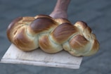 A freshly baked, golden-brown braided loaf of bread resting on a wooden paddle. The bread's surface has a shiny glaze, with perfectly intertwined strands that highlight its intricate design.