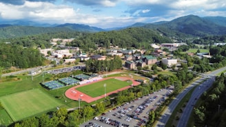 Aerial view of a sprawling college campus surrounded by lush green hills and mountains in the background. Central features include athletic fields with a running track, multiple buildings with varied architectural designs, and well-maintained lawns. A parking lot filled with cars lines the lower part of the scene, adjacent to a main road. The weather is clear, with a few clouds scattered in the blue sky, casting soft shadows over the landscape.
