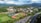 Aerial view of a sprawling college campus surrounded by lush green hills and mountains in the background. Central features include athletic fields with a running track, multiple buildings with varied architectural designs, and well-maintained lawns. A parking lot filled with cars lines the lower part of the scene, adjacent to a main road. The weather is clear, with a few clouds scattered in the blue sky, casting soft shadows over the landscape.