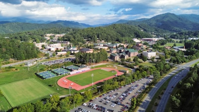 Aerial view of a sprawling college campus surrounded by lush green hills and mountains in the background. Central features include athletic fields with a running track, multiple buildings with varied architectural designs, and well-maintained lawns. A parking lot filled with cars lines the lower part of the scene, adjacent to a main road. The weather is clear, with a few clouds scattered in the blue sky, casting soft shadows over the landscape.