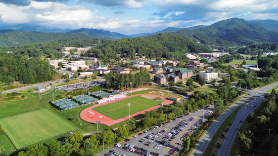 Aerial view of a sprawling college campus surrounded by lush green hills and mountains in the background. Central features include athletic fields with a running track, multiple buildings with varied architectural designs, and well-maintained lawns. A parking lot filled with cars lines the lower part of the scene, adjacent to a main road. The weather is clear, with a few clouds scattered in the blue sky, casting soft shadows over the landscape.