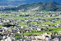 A sprawling rural landscape features numerous small homes and buildings scattered across lush green fields. In the background, gently sloping hills covered with dense forest further enhance the scenic environment. The picture captures the harmony between human habitation and nature.