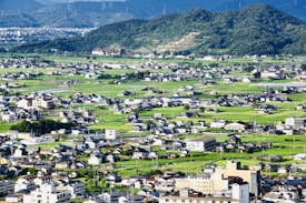 A sprawling rural landscape features numerous small homes and buildings scattered across lush green fields. In the background, gently sloping hills covered with dense forest further enhance the scenic environment. The picture captures the harmony between human habitation and nature.