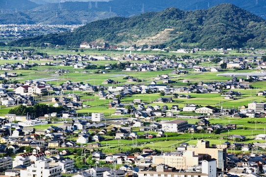A sprawling rural landscape features numerous small homes and buildings scattered across lush green fields. In the background, gently sloping hills covered with dense forest further enhance the scenic environment. The picture captures the harmony between human habitation and nature.