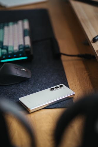 A smartphone with a white or light silver color rests on a dark desk mat beside a computer mouse. In the background, a mechanical keyboard with colorful keycaps and backlighting is visible on the wooden desk.