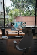 A home office setup with a curved monitor displaying an urban cityscape, complete with tall buildings and a tram on the street. A stylish, ergonomic chair is placed facing the desk. Surrounding the monitor are desktop speakers and a variety of tech accessories, including a keyboard and a tablet. Behind the setup, large windows reveal a view of lush greenery and a wooden fence.