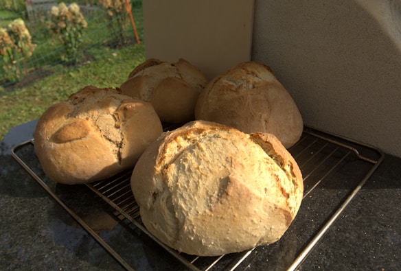 Four freshly baked loaves of bread are cooling on a metal rack placed on a dark kitchen counter. The loaves have a golden brown crust and are set against a backdrop of a garden, which is slightly out of focus in the background.