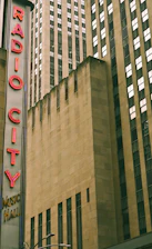 Tall buildings with beige exteriors dominate the image. A large, vertical sign with red neon letters spells out 'Radio City Music Hall' on the left side.