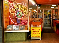 An entrance to a vintage record store features a large neon sign shaped like a record, surrounded by ads for vinyl, t-shirts, vintage clothing, and posters. A colorful array of vinyl records is displayed prominently at the storefront. There's a sign offering cash for used vinyl records. Inside, customers browse among racks filled with records.