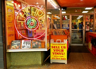 An entrance to a vintage record store features a large neon sign shaped like a record, surrounded by ads for vinyl, t-shirts, vintage clothing, and posters. A colorful array of vinyl records is displayed prominently at the storefront. There's a sign offering cash for used vinyl records. Inside, customers browse among racks filled with records.