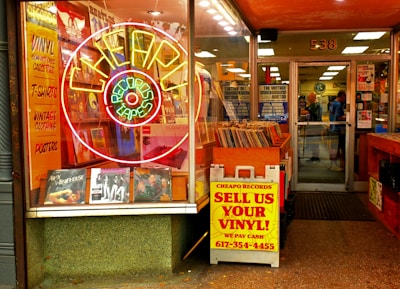 An entrance to a vintage record store features a large neon sign shaped like a record, surrounded by ads for vinyl, t-shirts, vintage clothing, and posters. A colorful array of vinyl records is displayed prominently at the storefront. There's a sign offering cash for used vinyl records. Inside, customers browse among racks filled with records.