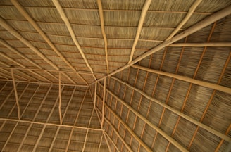 A close-up view of a thatched roof structure, featuring interwoven natural materials like wooden beams and dried palm leaves. The pattern of the roof showcases geometric lines and angles, creating a sense of texture and craftsmanship.