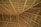 A close-up view of a thatched roof structure, featuring interwoven natural materials like wooden beams and dried palm leaves. The pattern of the roof showcases geometric lines and angles, creating a sense of texture and craftsmanship.