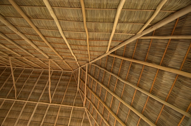 A close-up view of a thatched roof structure, featuring interwoven natural materials like wooden beams and dried palm leaves. The pattern of the roof showcases geometric lines and angles, creating a sense of texture and craftsmanship.