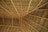 A close-up view of a thatched roof structure, featuring interwoven natural materials like wooden beams and dried palm leaves. The pattern of the roof showcases geometric lines and angles, creating a sense of texture and craftsmanship.